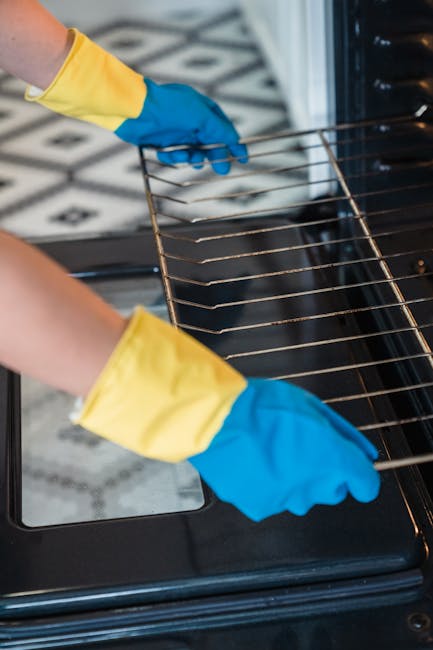 A person wearing yellow and blue rubber cleaning gloves is placing a metal oven rack into a black oven, which is part of a modern kitchen with a tiled black and white patterned floor. The oven interior appears clean and free of food residue, with the door open to reveal the shiny oven cavity. Natural lighting enhances the clarity of the surfaces, emphasizing the thoroughness of the oven cleaning process. This image showcases the type of surface and deep cleaning services provided by Oven Cleaning Kennington, highlighting the importance of proper maintenance and sanitisation of domestic appliances for hygiene and functionality.