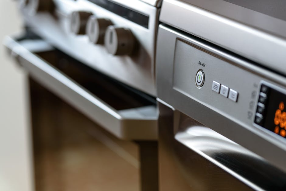 Close-up view of a stainless steel oven with a digital control panel and a nearby gas hob with three knobs, situated in a modern kitchen. The oven's surface appears clean and well-maintained, with a smooth metallic finish reflecting light. The control buttons on the oven include an on/off switch and preset options. The surrounding kitchen area features a countertop and part of a wooden floor, highlighting the space's tidy and hygienic condition. The image emphasizes surface cleaning and maintenance as provided by Oven Cleaning Kennington, specializing in domestic cleaning and deep oven cleaning services.