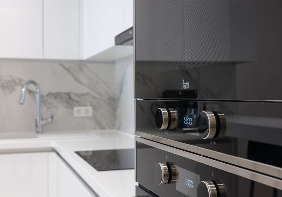 A person wearing yellow and blue rubber cleaning gloves is placing a metal oven rack into a black oven, which is part of a modern kitchen with a tiled black and white patterned floor. The oven interior appears clean and free of food residue, with the door open to reveal the shiny oven cavity. Natural lighting enhances the clarity of the surfaces, emphasizing the thoroughness of the oven cleaning process. This image showcases the type of surface and deep cleaning services provided by Oven Cleaning Kennington, highlighting the importance of proper maintenance and sanitisation of domestic appliances for hygiene and functionality.