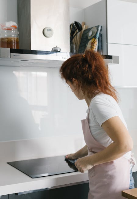 A woman with long, curly reddish-brown hair wearing a white t-shirt and light pink apron is engaged in surface cleaning in a modern kitchen. She is using a black cloth to wipe a sleek, black glass induction cooktop situated on a white countertop. The kitchen features white cabinets, some with a glossy finish, and a metallic range hood above the cooktop. Visible in the background are a jar of cleaning supplies, a thermostat on the wall, and a few cookbooks placed upright. The lighting is bright and natural, highlighting the clean and tidy surface of the workspace, which shows no signs of dust, grease, or stains. This scene represents professional deep cleaning and sanitisation services provided by Oven Cleaning Kennington, focused on maintaining hygiene and cleanliness in residential kitchens near Kennington Park SE11.