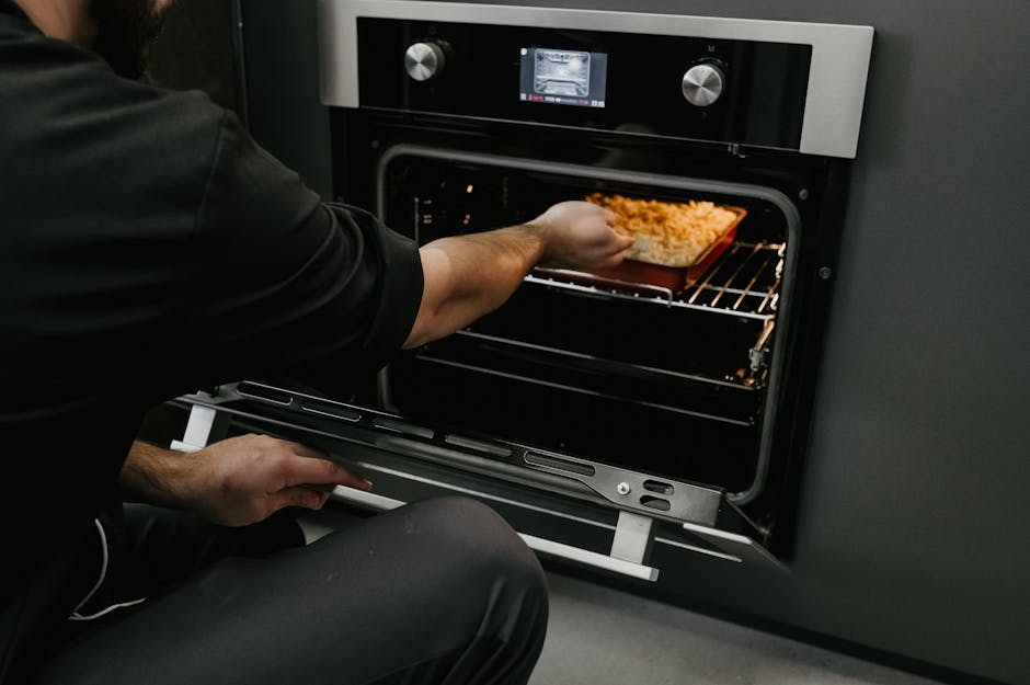 A person wearing a white shirt and yellow and blue cleaning gloves is cleaning the interior of an oven with a wire rack, inside a kitchen with a ceramic tiled floor featuring a black and white pattern. The oven door is open, revealing its clean, dark interior with oven lights illuminating the inside. The exterior of the oven has a white finish with control knobs on the front. This image exemplifies thorough surface cleaning and deep cleaning processes, associated with domestic cleaning services offered by Oven Cleaning Kennington, focused on maintaining hygiene and appliance sanitisation in residential kitchens.