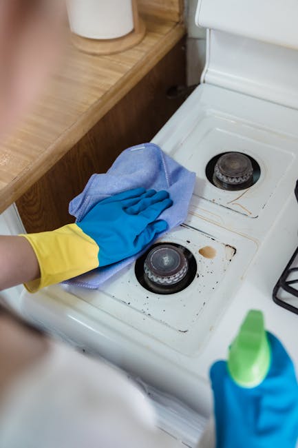 A person wearing yellow and blue rubber gloves is cleaning the interior of a white electric oven with a wire rack inside. The oven's glass door is open, revealing the illuminated dark interior. The ki