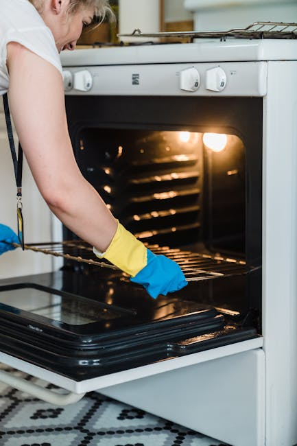 A person wearing yellow and blue cleaning gloves is inserting a wire rack into an open oven for deep cleaning. The oven has a white exterior with control knobs on the front panel and a black glass door with a reflective surface. Inside, the oven's interior is illuminated, revealing clean metal racks. The surrounding kitchen area features light-colored wooden cabinets and a patterned black-and-white tiled floor. Natural light illuminates the scene, emphasizing the cleanliness of the surfaces. This image, associated with Oven Cleaning Kennington, depicts surface cleaning and maintenance in a domestic kitchen setting to prevent hidden costs in oven cleaning services.