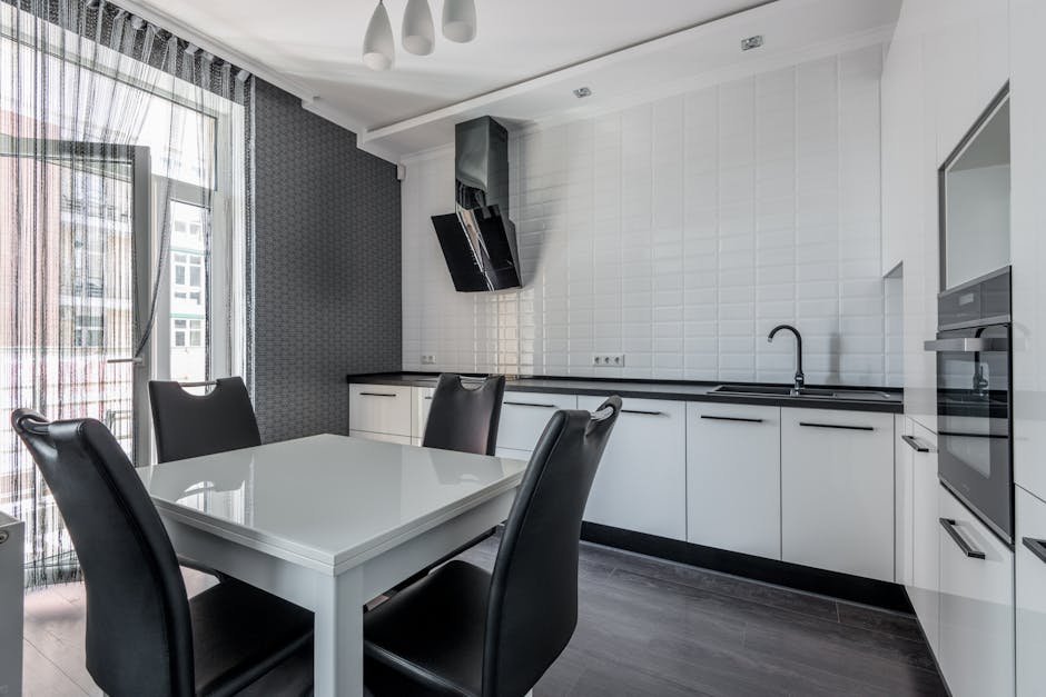 A modern, well-lit kitchen with white cabinetry and black countertops, featuring a stainless steel sink with a black faucet on the right side. The room has a tiled backsplash with a subtle geometric pattern, and a black range hood is mounted above the stovetop. A white rectangular dining table with a glossy finish is centrally positioned, surrounded by four black leather chairs with high backs. The kitchen floor appears to be dark wood or laminate, and large glass doors with sheer curtains on the left allow natural light to flood in, enhancing the clean and tidy appearance. There are no visible stains or clutter, and the surfaces, including the countertops and table, appear spotless. This setting exemplifies comprehensive surface cleaning and sanitisation in a domestic kitchen environment, undertaken by Oven Cleaning Kennington as part of their expert cleaning service for SE11 flats, referenced in the Landlord oven cleaning checklist for Kenton, KENNINGTON.