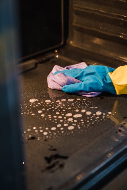A close-up view of a black oven surface being cleaned, with a person wearing bright yellow and blue gloves using a cloth to wipe away soap suds and residue. The oven interior is made of stainless steel, and the surface shows signs of recent cleaning, with foam and water droplets visible. The scene is illuminated by indoor lighting, highlighting the shine and cleanliness of the oven. The image illustrates a step in surface cleaning and maintenance, relevant to domestic oven deep cleaning services offered by Oven Cleaning Kennington, aligned with the landlord oven cleaning checklist for SE11 flats in Kennington.