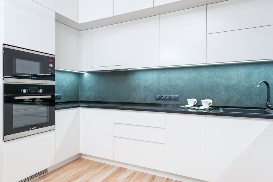 A modern, well-lit kitchen with white cabinetry and black countertops, featuring a stainless steel sink with a black faucet on the right side. The room has a tiled backsplash with a subtle geometric pattern, and a black range hood is mounted above the stovetop. A white rectangular dining table with a glossy finish is centrally positioned, surrounded by four black leather chairs with high backs. The kitchen floor appears to be dark wood or laminate, and large glass doors with sheer curtains on the left allow natural light to flood in, enhancing the clean and tidy appearance. There are no visible stains or clutter, and the surfaces, including the countertops and table, appear spotless. This setting exemplifies comprehensive surface cleaning and sanitisation in a domestic kitchen environment, undertaken by Oven Cleaning Kennington as part of their expert cleaning service for SE11 flats, referenced in the Landlord oven cleaning checklist for Kenton, KENNINGTON.