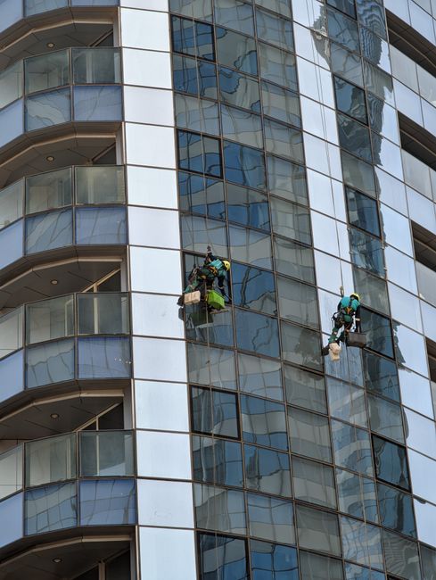 A worker wearing a yellow safety helmet and dark clothing is suspended by ropes inside a building, using a high-pressure water spray to clean large, vertical glass panels with a reflective surface. Th