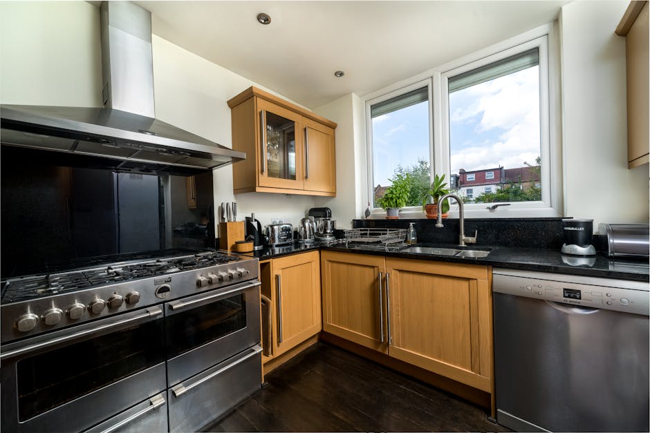 A modern residential kitchen featuring a stainless steel range cooker with multiple burners and a black backsplash, adjacent to honey-colored wooden cabinetry with glass-paneled upper doors and sleek metal handles. The black granite countertop holds various small appliances, including a kettle, toaster, and coffee machine, with a drying rack near the sink. The sink, with a silver faucet, is positioned beneath a large double-pane window that allows natural light to brighten the room and offers a view of neighboring houses and greenery outside. A potted plant on the windowsill adds a touch of green. The kitchen appears clean and well-maintained, with shiny surfaces and no visible clutter, reflecting professional domestic cleaning consistent with surface cleaning, hygiene, and maintenance standards by Oven Cleaning Kennington.