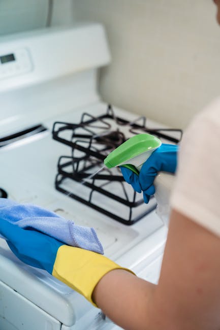A person wearing blue and yellow protective gloves is performing surface cleaning with a spray bottle and a cloth in a kitchen setting. The background features a white gas stove with black metal burners and a beige wall. The surface around the stove appears clean and well-maintained, indicating a thorough cleaning process. The lighting is natural and bright, highlighting the shiny, sanitized surfaces. This image illustrates domestic surface cleaning, deep cleaning, or sanitisation in a residential kitchen, aligning with the services offered by Oven Cleaning Kennington, as seen on the page for same day oven cleaning for Oval residents.