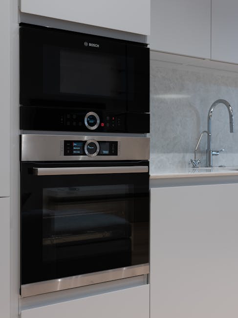 A modern residential kitchen featuring a stainless steel range cooker with multiple burners and a black backsplash, adjacent to honey-colored wooden cabinetry with glass-paneled upper doors and sleek metal handles. The black granite countertop holds various small appliances, including a kettle, toaster, and coffee machine, with a drying rack near the sink. The sink, with a silver faucet, is positioned beneath a large double-pane window that allows natural light to brighten the room and offers a view of neighboring houses and greenery outside. A potted plant on the windowsill adds a touch of green. The kitchen appears clean and well-maintained, with shiny surfaces and no visible clutter, reflecting professional domestic cleaning consistent with surface cleaning, hygiene, and maintenance standards by Oven Cleaning Kennington.